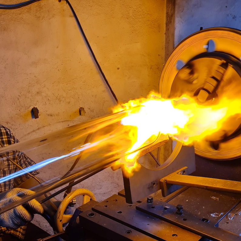 May include: A glassblowing process in action, with a long, clear glass tube being heated by a bright orange flame. The flame is intense, and the glass is being shaped with tools. The background is a workshop setting.