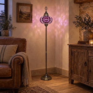 May include: A tall floor lamp featuring a round, mosaic glass shade in shades of purple and white. The lamp has a slender, dark metal stand with a circular base and an ornate metal finial at the top, casting a patterned light on the wall.