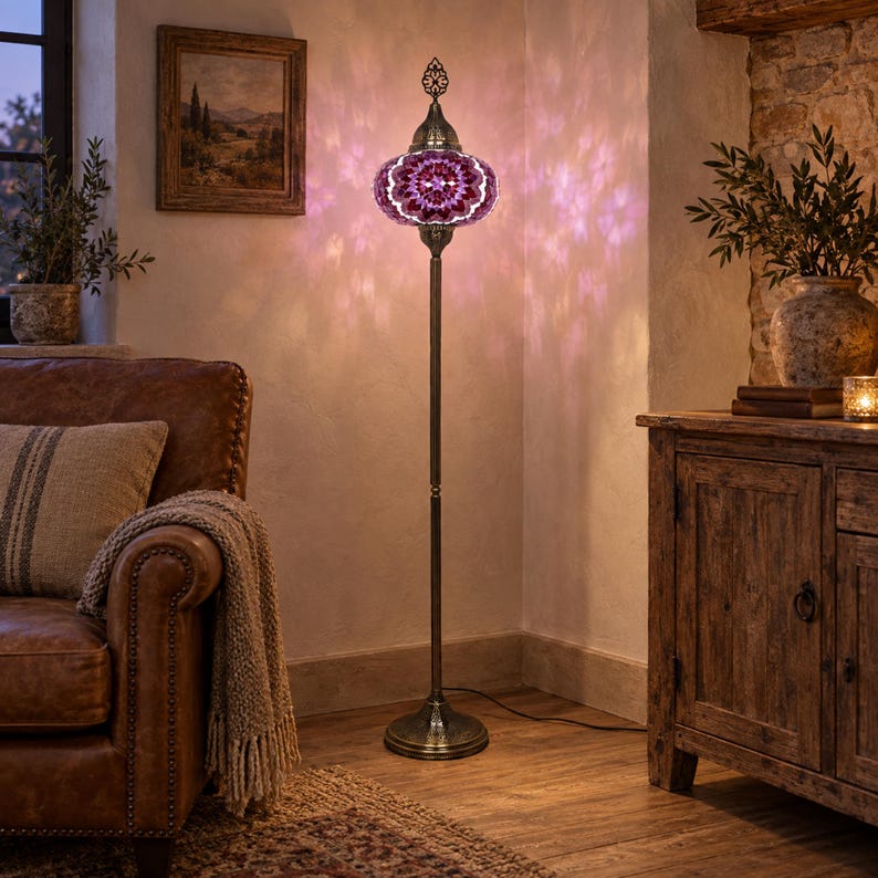 May include: A tall floor lamp featuring a round, mosaic glass shade in shades of purple and white. The lamp has a slender, dark metal stand with a circular base and an ornate metal finial at the top, casting a patterned light on the wall.