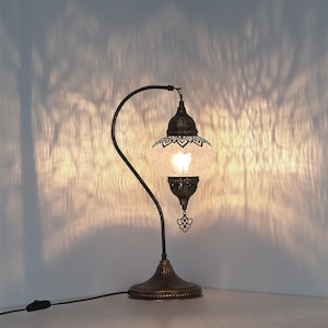 May include: A decorative table lamp with a bronze finish. The lamp features a curved arm supporting a globe-shaped shade with intricate cut-out patterns. The shade emits a warm light, casting decorative shadows on the wall. The lamp has a detailed base and a dangling ornament.