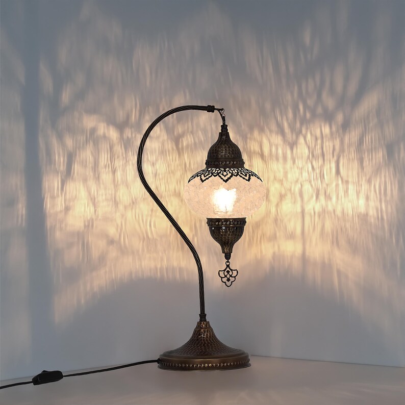 May include: A decorative table lamp with a bronze finish. The lamp features a curved arm supporting a globe-shaped shade with intricate cut-out patterns. The shade emits a warm light, casting decorative shadows on the wall. The lamp has a detailed base and a dangling ornament.
