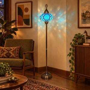 May include: A tall floor lamp featuring a mosaic glass shade in shades of blue and white with a star pattern. The lamp has a slender, dark metal stand and a circular base, casting a patterned light projection onto the surrounding wall surface.