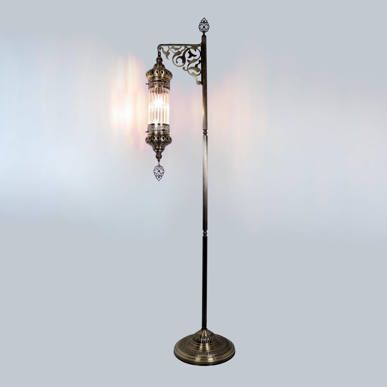 May include: A bronze-colored floor lamp with an ornate design. The lamp features a cylindrical glass shade with vertical lines, suspended from a decorative arm. The base and top of the lamp have intricate patterns, and the lamp stands on a circular base.