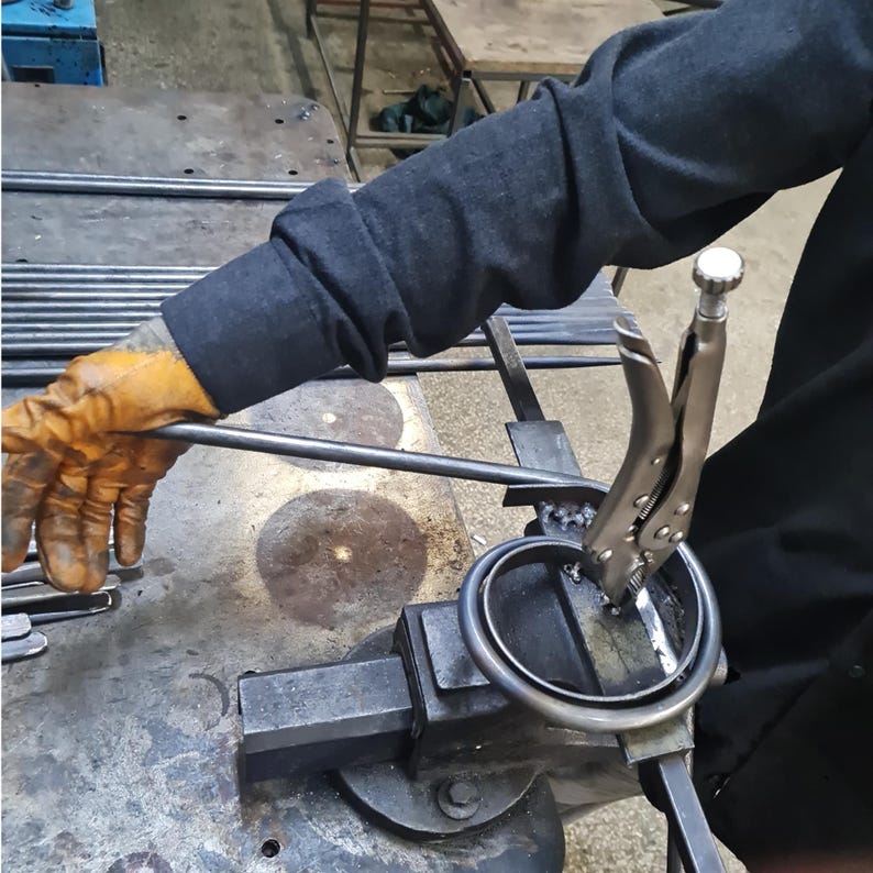 May include: A metalworker uses pliers to bend a metal rod into a circular shape. The worker wears a dark gray long-sleeved shirt and yellow work gloves. The metal rod is held in a vise.