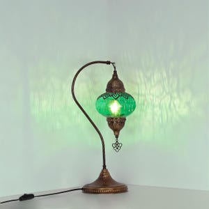 May include: A decorative table lamp with a green glass shade and a bronze-colored metal base and curved arm. The shade has a patterned design and emits a warm glow. The lamp is plugged into a wall outlet.