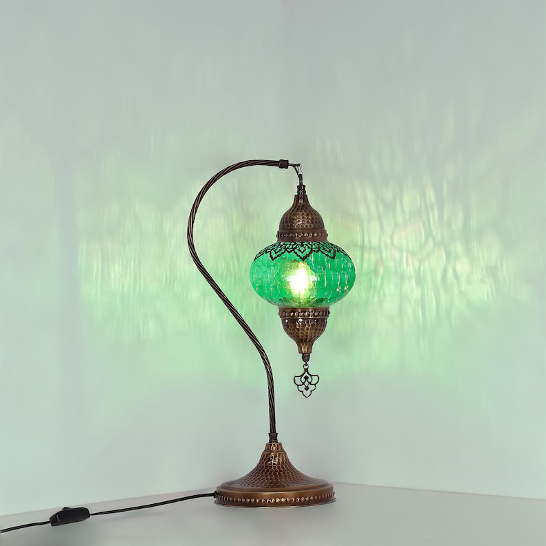 May include: A decorative table lamp with a green glass shade and a bronze-colored metal base and curved arm. The shade has a patterned design and emits a warm glow. The lamp is plugged into a wall outlet.