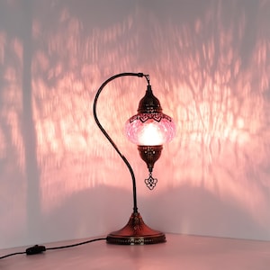 May include: A decorative table lamp with a pink glass shade and a dark bronze metal base. The lamp has an ornate design with a curved arm and a decorative pendant. The lamp is plugged into a wall outlet.