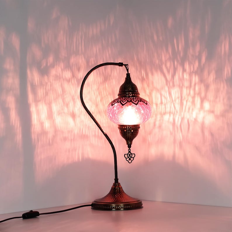 May include: A decorative table lamp with a pink glass shade and a dark bronze metal base. The lamp has an ornate design with a curved arm and a decorative pendant. The lamp is plugged into a wall outlet.