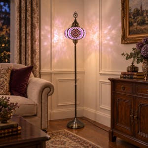 May include: A floor lamp featuring a mosaic glass shade with pink, white, and black geometric patterns. The lamp has a slender, dark metal stand and a circular base, emitting a soft, patterned light that casts shadows on the surrounding wall.
