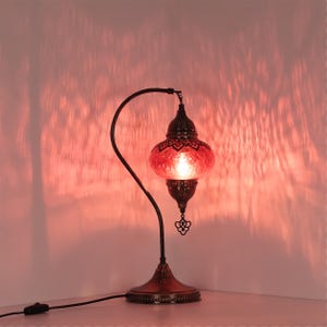 May include: A decorative table lamp with a red glass shade and a dark metal base. The lamp has an ornate design and casts a warm, reddish glow. The lamp's curved arm and intricate details add to its aesthetic appeal.