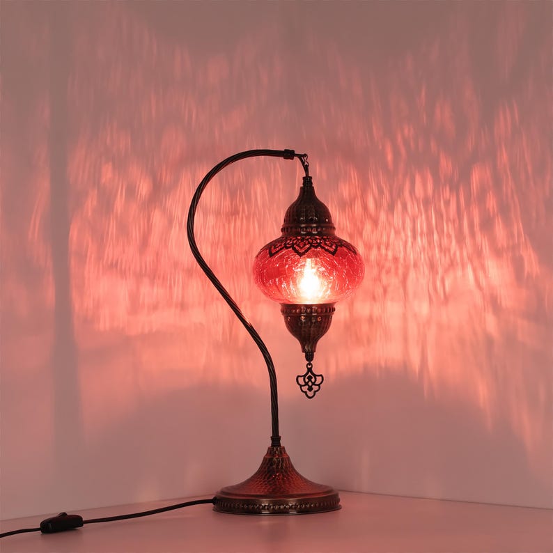 May include: A decorative table lamp with a red glass shade and a dark metal base. The lamp has an ornate design and casts a warm, reddish glow. The lamp's curved arm and intricate details add to its aesthetic appeal.