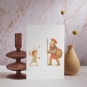 May include: A greeting card featuring an illustration of David and Goliath. The artwork depicts a young boy with a slingshot facing a large warrior holding a spear and shield. A brown vase with yellow flowers and a decorative glass candle holder are also in the image.