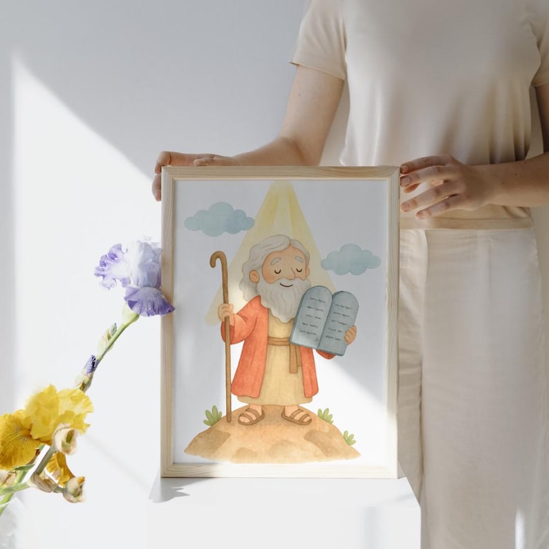 May include: A framed watercolor print featuring a cartoon illustration of a biblical figure with a long white beard, holding a staff and tablets. The figure is wearing a red robe and standing on a small hill with clouds and a yellow light in the background. The frame is made of light wood.