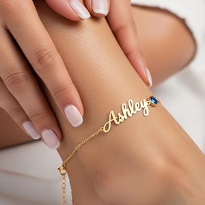 May include: A gold-colored anklet with the name "Ashley" in cursive script. The anklet features a small blue gemstone and is displayed on a person's ankle. The background is a soft, neutral color.