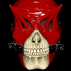 May include: A glossy red mask with a skull-like lower face and a horned upper section. The mask has eye openings and a strap with metal buckles. The skull portion is off-white with detailed teeth.