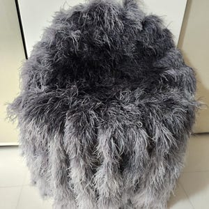 Könnte beinhalten: Ein runder, grauer, flauschiger Hocker mit Farbverlauf. Der Hocker ist mit langem, weichem, federartigem Material bedeckt, das von Dunkelgrau oben zu einem helleren Grau unten übergeht. Das Design ist modern und luxuriös.