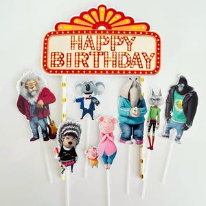 May include: A colorful cake topper featuring characters from the animated movie "Sing." The topper includes a marquee sign that reads "HAPPY BIRTHDAY" and several character cutouts on sticks, including a lion, koala, elephant, pig, and gorilla.