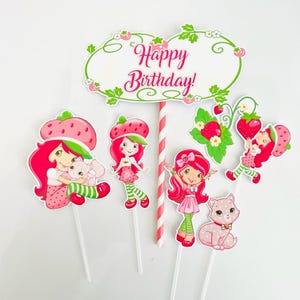 May include: A collection of colorful birthday cake toppers featuring a strawberry theme. The toppers include cartoon characters, strawberries, and a sign that reads "Happy Birthday!" in pink script. A pink and white striped straw is also included.