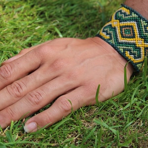 Peut inclure: Un bracelet tissé aux couleurs vert, jaune et noir, orné d'un motif géométrique. Le bracelet est porté sur un poignet, posé sur de l'herbe verte. Le design du bracelet est complexe et attrayant.