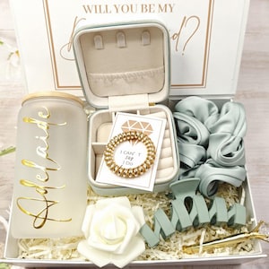 May include: A bridesmaid gift box containing a frosted glass tumbler with "Bride" in gold lettering, a small jewellery case, a gold coil bracelet, and a hair claw clip. The box also includes a white rose and a pen with a diamond-shaped top.