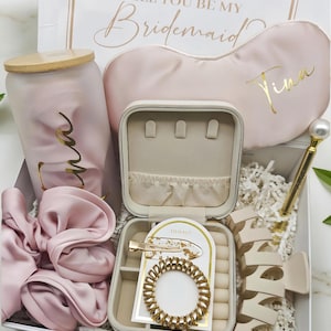 Könnte beinhalten: Eine Geschenkbox mit verschiedenen Artikeln, darunter eine rosa Schlafmaske, ein Becher, ein Scrunchie, ein Schmuckkästchen, eine Haarspange, ein Spiralhaargummi und Holzbuchstaben. Die Box ist weiß mit goldenem Schriftzug "Willst du meine Brautjungfer sein?".
