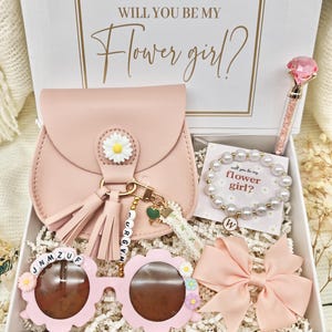 May include: A gift box with the text "WILL YOU BE MY Flower girl?" contains a pink purse, sunglasses, a bracelet, a pen, and a bow. The sunglasses are flower-shaped. The bracelet has the text "will you be my flower girl?"