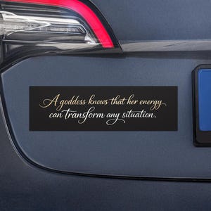 May include: A black rectangular car magnet with the text "A goddess knows that her energy can transform any situation" in gold script. The magnet is affixed to the rear of a dark gray car.