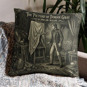 Può includere: Cuscino decorativo con un'illustrazione in bianco e nero da "Il ritratto di Dorian Gray" di Oscar Wilde. Il design raffigura un uomo in una stanza con un dipinto, teschi e elementi gotici. Il cuscino è posto su una coperta fantasia.