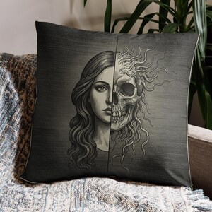 Könnte beinhalten: Ein dekoratives Zierkissen mit geteiltem Design. Eine Seite zeigt das Gesicht einer Frau mit langen, welligen Haaren, die andere einen menschlichen Schädel. Das Kissen ist grau mit strukturiertem Aussehen und liegt auf einem grauen Sofa mit einer blau-weißen Decke.