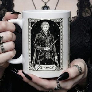 Puede incluir: Taza de cerámica blanca con una ilustración en blanco y negro de un personaje sosteniendo un cuchillo, con el nombre "Astarion" debajo. La taza tiene un asa y la sostiene una persona con esmalte de uñas negro y varios anillos.