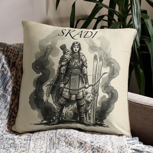 Puede incluir: Un cojín crema con una ilustración en blanco y negro de una figura con arco y flechas, esquís y un lobo. La palabra "SKADI" está impresa encima de la figura. El cojín está sobre una manta estampada.