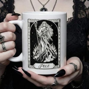 May include: White ceramic mug featuring a black and white illustration of a woman with long hair, holding a staff, and surrounded by flames. The word "PELE" is printed at the bottom of the image. The mug has a white handle.