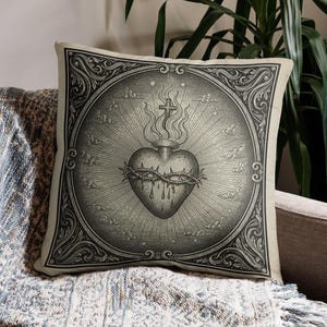 May include: Square cushion featuring a detailed black and white illustration of a heart encircled by a crown of thorns, a cross, and flames. The design is set within a decorative frame.