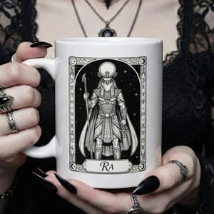 Può includere: Tazza in ceramica bianca con un'illustrazione in bianco e nero del dio egizio Ra. Il disegno include geroglifici e la parola "Ra". La tazza ha un manico bianco ed è tenuta da una persona.