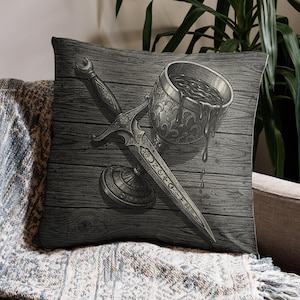 May include: Decorative throw pillow featuring a detailed illustration of a dagger and chalice with liquid spilling out. The design is in grayscale, set against a wood-grain background. The pillow is placed on a sofa with a patterned throw.