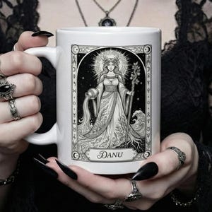 Puede incluir: Taza de cerámica blanca con una ilustración en blanco y negro de una mujer coronada sosteniendo una jarra y un bastón, con el nombre "DANU" debajo. El diseño está enmarcado con bordes decorativos y elementos celestiales.
