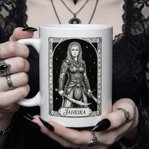 May include: White ceramic mug featuring a black and white illustration of a woman warrior holding two swords, with the name "Jaheira" below. The design is framed with decorative elements. The mug is held by a person with long, black fingernails.