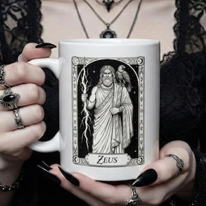 Op de afbeelding: Witte keramische mok met een zwart-wit illustratie van Zeus, die een bliksemschicht vasthoudt en met een adelaar op zijn schouder. Het woord "Zeus" staat eronder gedrukt. De mok heeft een standaard handvat.