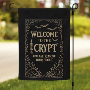 Könnte beinhalten: Schwarze Gartenflagge mit dem Text "WELCOME TO THE CRYPT (PLEASE REMOVE YOUR SHOES)". Die Flagge hat einen dekorativen Rand mit Fledermäusen und einer Kerze. Die Flagge hängt an einem schwarzen Metallständer.