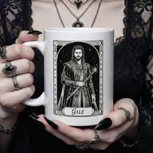 Puede incluir: Taza de cerámica blanca con una ilustración en blanco y negro de un hombre sosteniendo un bastón, con el nombre "Gale" debajo. El diseño está enmarcado con elementos decorativos. La taza es sostenida por una persona con esmalte de uñas negro y varios anillos.