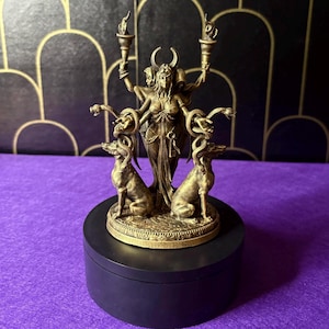 Op de afbeelding: Een bronzen beeld van een godin met drie gezichten, die fakkels vasthoudt, geflankeerd door twee honden en slangen. Het beeld is gemonteerd op een zwarte, ronde basis. De achtergrond is donker met een gouden patroon.