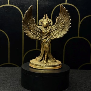 Könnte beinhalten: Eine goldfarbene Statue einer geflügelten Gottheit mit erhobenen Armen, die auf einem kreisförmigen Sockel steht. Die Statue steht auf einem schwarzen, runden Sockel vor einem dunklen Hintergrund mit goldenen Bogenmustern. Die Flügel der Statue sind weit ausgebreitet.