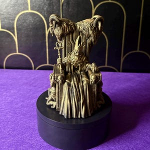 May include: A gold-colored figurine of a robed figure seated on a throne, holding a staff with an ankh. The throne features ram heads and skull details. The figurine sits on a black, round base, with a purple surface in the background.