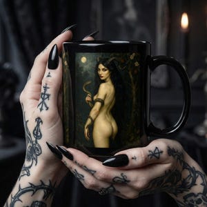 Op de afbeelding: Zwarte keramische mok met een schilderij van een naakte vrouw met lang zwart haar, hoorns en een slang. Het kunstwerk speelt zich af in een bos met volle maan. De mok heeft een zwart handvat.