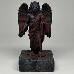 May include: A detailed sculpture of a lion-headed figure with large wings, draped in red fabric. The statue stands on a textured, dark gray base. The lion's face is fierce, and the wings are a deep red and black.