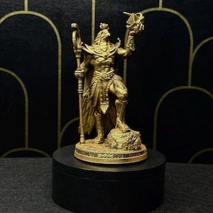May include: A gold-colored statue of a figure with a bird head, holding a staff and a small pyramid. The statue is standing on a rock base and is displayed on a black pedestal. The background is black with gold arched patterns.