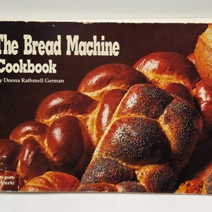 May include: The cover of "The Bread Machine Cookbook" by Donna Rathmell German. The book features a dark brown cover with the title in white. The image shows various types of freshly baked bread, including loaves with poppy seeds.