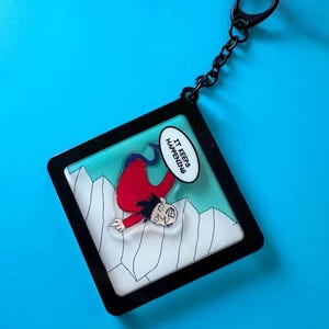 Puede incluir: Un llavero negro con un gráfico de dibujos animados de una persona cayendo. La persona lleva una camisa roja y se cae por una montaña. Un bocadillo dice "IT KEEPS HAPPENING".