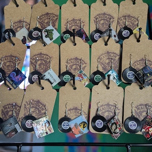 May include: A display of handmade earrings featuring miniature vinyl records and album art. Each pair hangs from a brown tag with the word "MAUDLIN" printed on it. The earrings showcase various album covers and designs, offering a unique, music-themed accessory.
