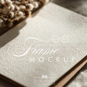 Könnte beinhalten: Ein Rahmen-Mockup mit dem Text "Frame Mockup" in einer Schreibschrift. Der Rahmen ist offen, mit einer strukturierten, cremefarbenen Oberfläche. Getrocknete Blumen befinden sich in der oberen linken Ecke. Der Rahmen hat den Text "Moku Mocks".
